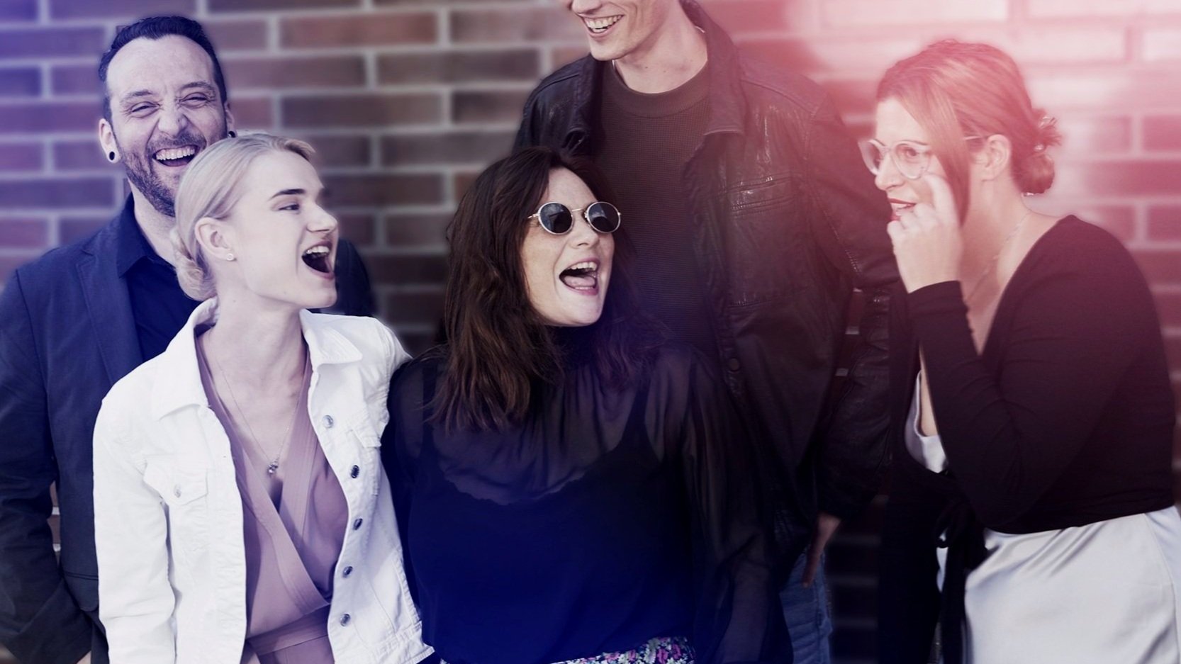 Group of five friends laughing and talking outdoors against a brick wall, with some sunlight creating a slight pinkish glow.