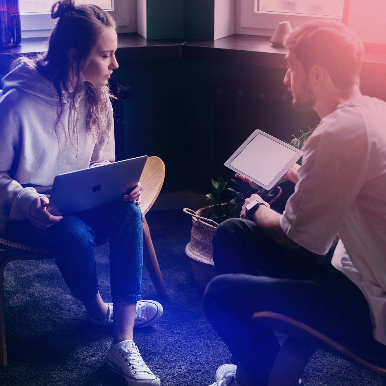 A woman and a man sitting and facing each other indoors, each holding a tablet device. The woman is seated on a wooden chair, and the man is sitting cross-legged on a cushioned chair, with a potted plant nearby.