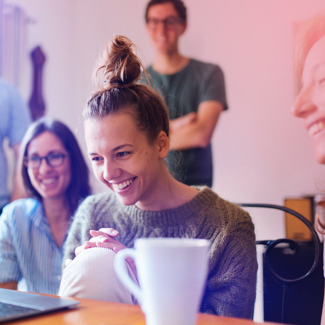 Group of four young adults enjoying a social moment indoors, smiling and engaging with each other around a table with a laptop and a white coffee mug.