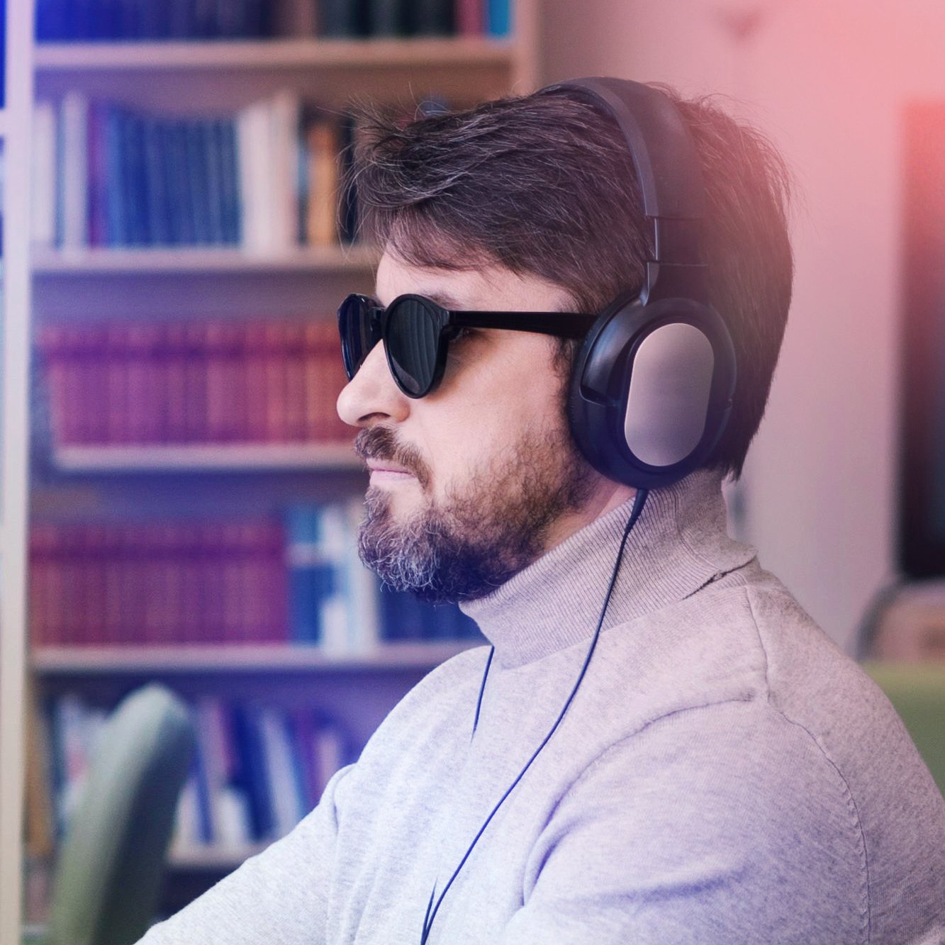 A man with dark hair and beard wearing sunglasses and over-ear headphones, sitting indoors with a bookshelf in the background.