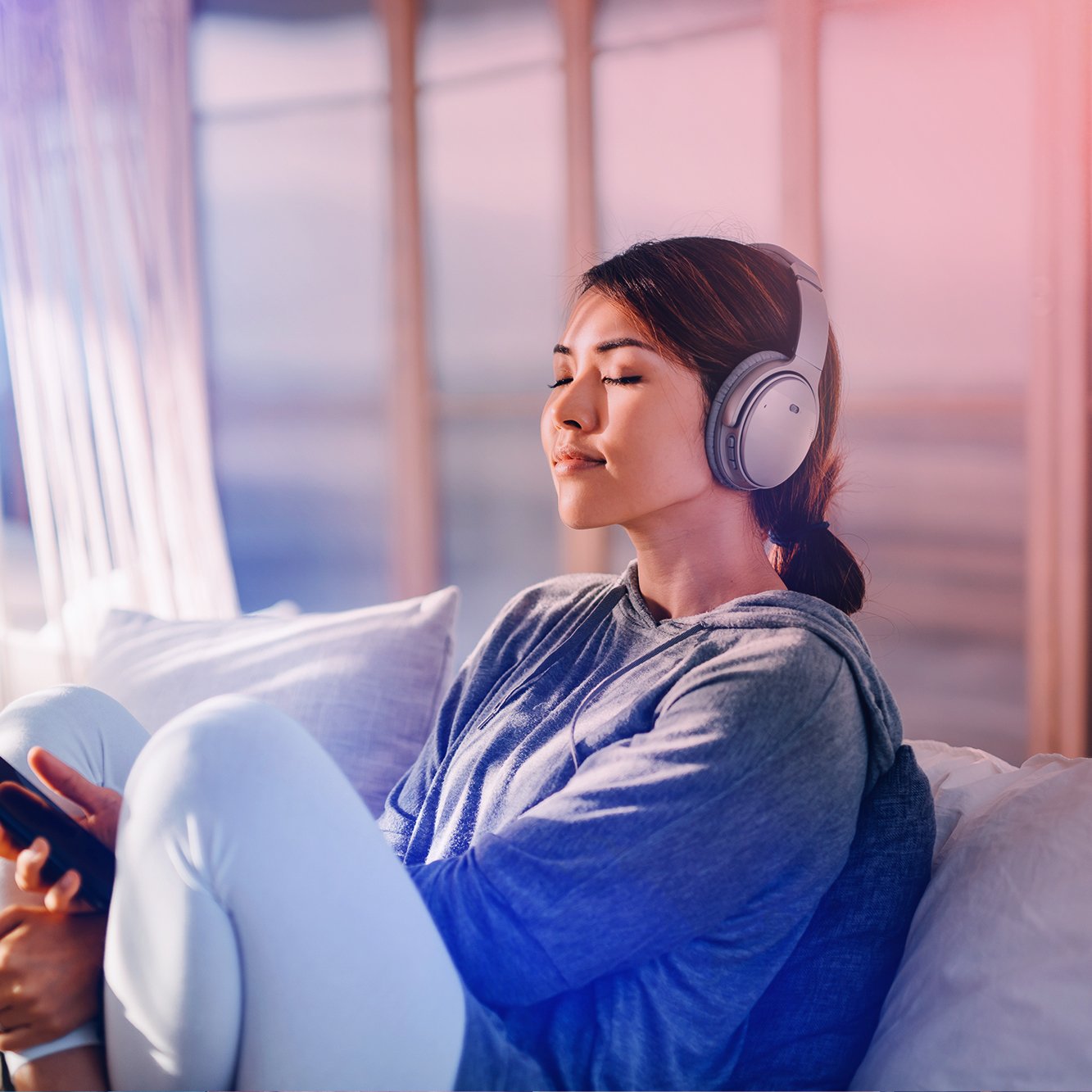 Young woman relaxing on a couch with eyes closed, wearing headphones and holding a smartphone, in a sunlit room.