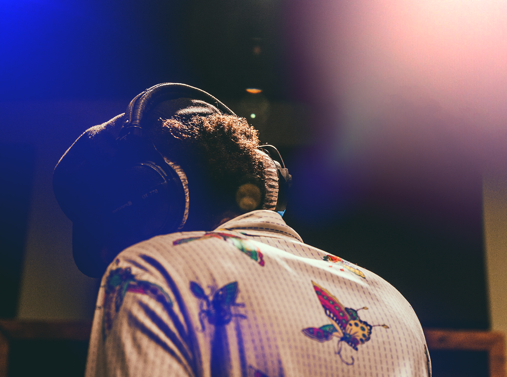 Back of a person wearing large headphones and a shirt with colorful butterfly patterns, with sunlight shining from the upper right corner.