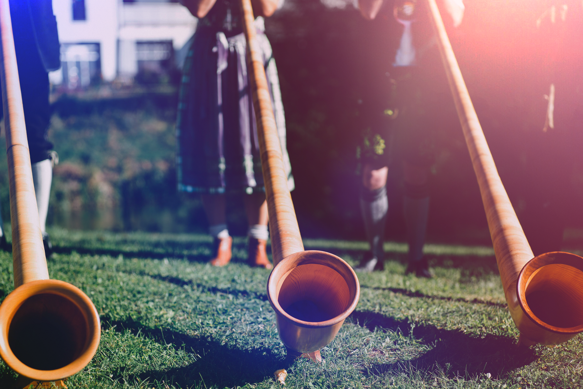 A group of people playing traditional outdoor wooden horn instruments called alphorns on grass with sunlight shining in the background.