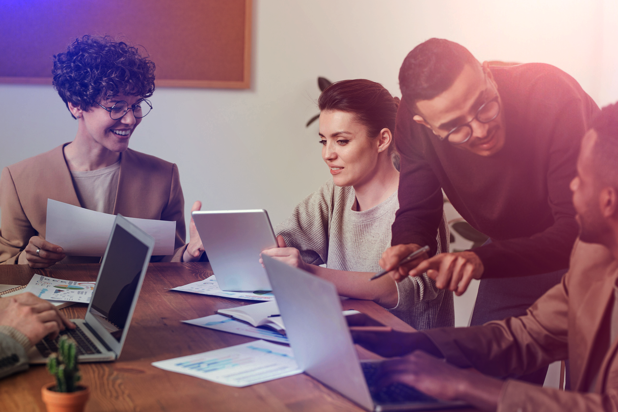 A group of five diverse professionals working together in a meeting room with laptops, papers, and tablets on the table.