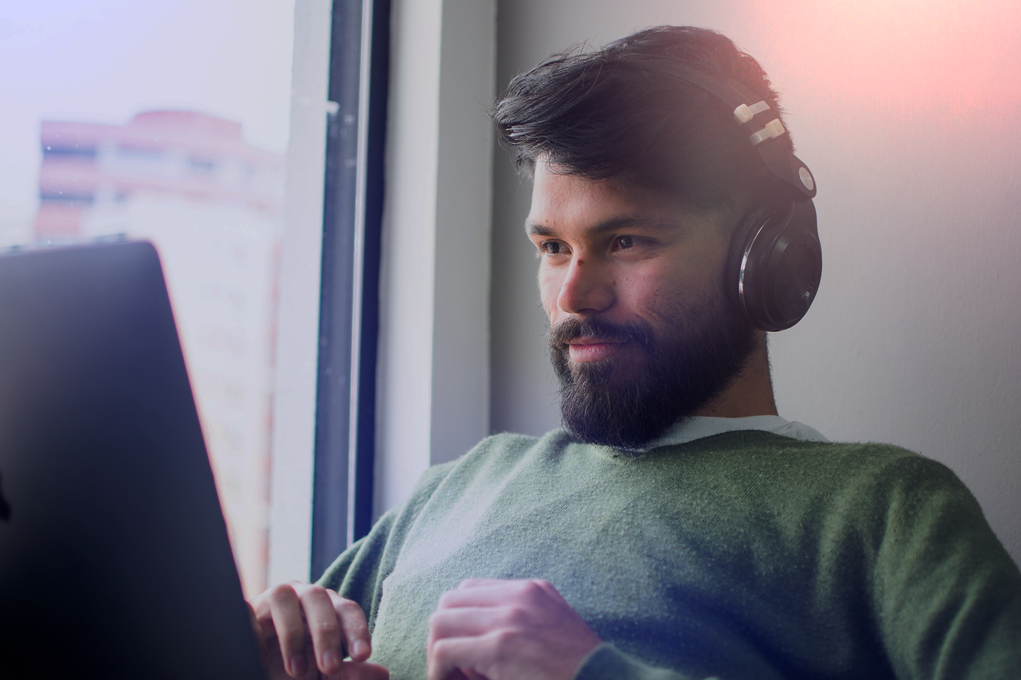 A man with dark hair and a beard wearing headphones, sitting by a window, looking at a laptop screen with a slight smile.