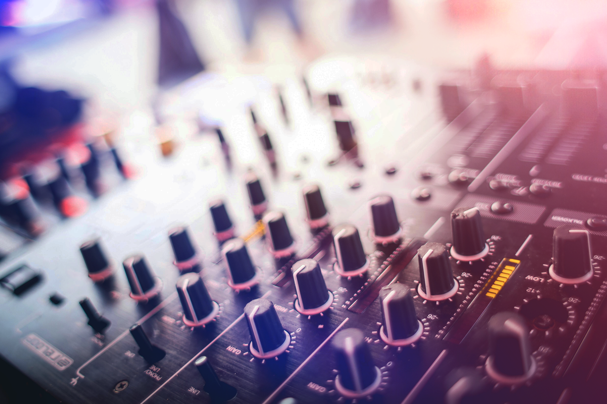 Close-up of a professional audio mixer with multiple knobs and sliders, blurred background, and a pinkish glow.