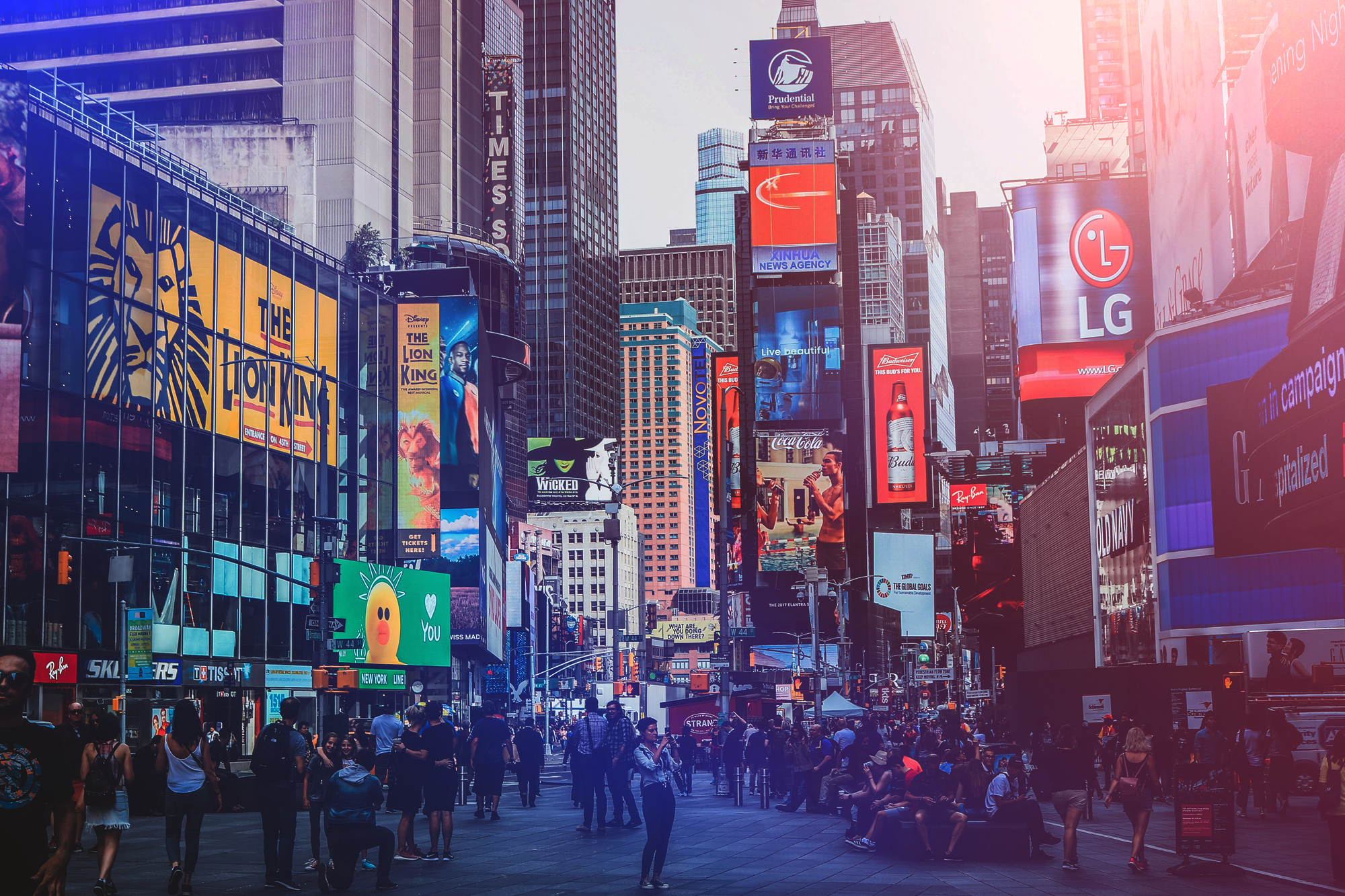 Crowded city street with large digital billboards and neon signs, including advertisements for The Lion King, Wicked, LG, Coca-Cola, and Ray-Ban, among tall buildings, with people walking and sitting.