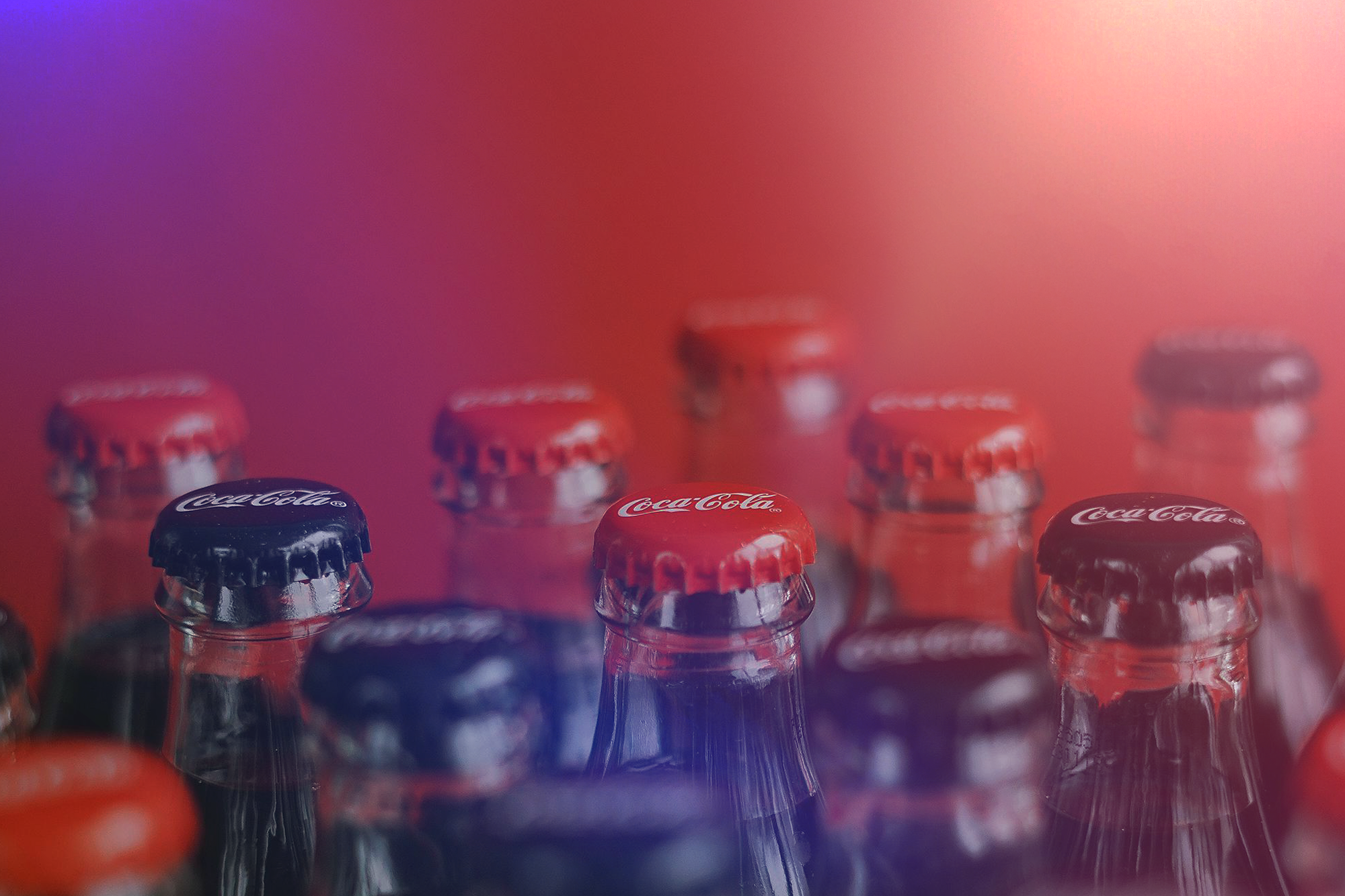 Close-up of various Coca-Cola glass bottles with red and black bottle caps against a colorful blurred background.