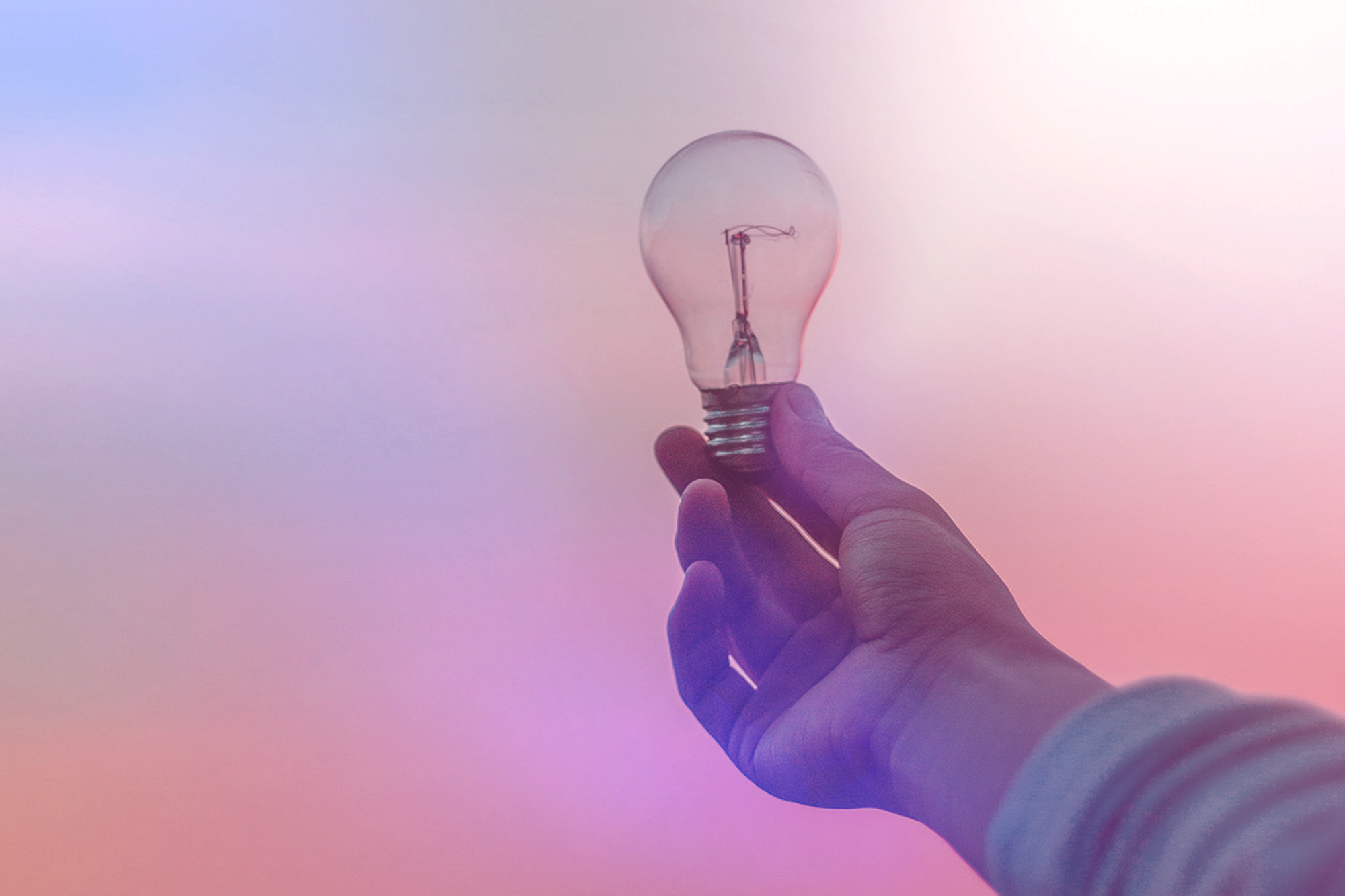 A hand holding a clear light bulb against a pastel gradient background